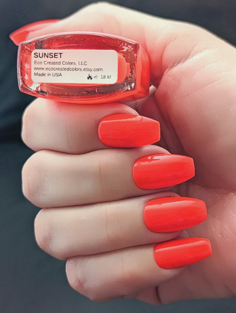 May include: A bottle of bright orange nail polish labeled "SUNSET" by Eco Created Colors, LLC. The bottle is in a hand with four orange nails painted with the polish.