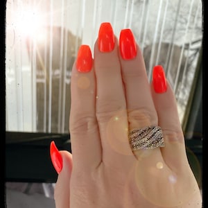 May include: A hand with bright orange nail polish and a silver ring with clear stones. The hand is in front of a window with sunlight shining through.