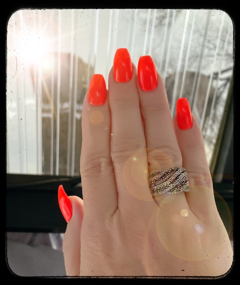 May include: A hand with bright orange nail polish and a silver ring with clear stones. The hand is in front of a window with sunlight shining through.