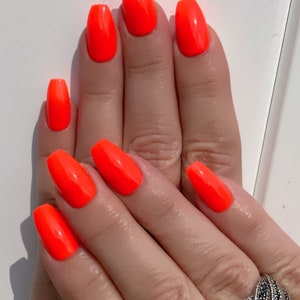 May include: Close-up of hands with bright orange nail polish on all ten fingernails. The nails are long and shaped into a coffin shape. The person is wearing a silver ring with black stones on their ring finger.