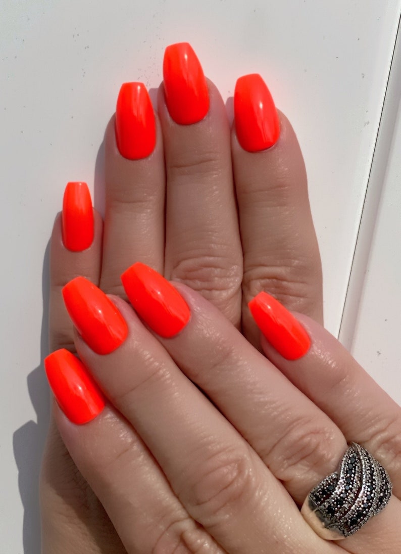 May include: Close-up of hands with bright orange nail polish on all ten fingernails. The nails are long and shaped into a coffin shape. The person is wearing a silver ring with black stones on their ring finger.