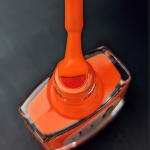 May include: A close-up of a bright orange nail polish bottle with a brush dipped in the polish. The bottle is made of clear glass and has a black label.
