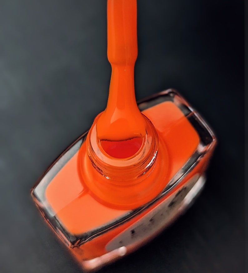 May include: A close-up of a bright orange nail polish bottle with a brush dipped in the polish. The bottle is made of clear glass and has a black label.