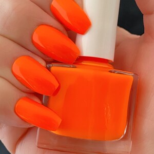 May include: A clear glass bottle of bright orange nail polish with a white cap. The bottle is held in a hand with several nails painted in the same bright orange color.
