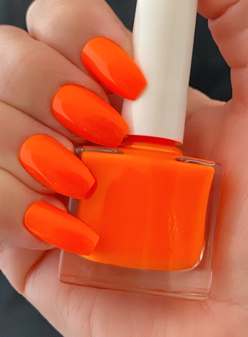 May include: A clear glass bottle of bright orange nail polish with a white cap. The bottle is held in a hand with several nails painted in the same bright orange color.