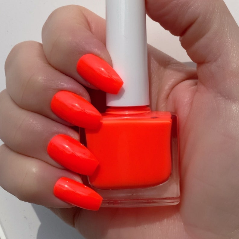 May include: A hand holding a bottle of bright orange nail polish. The bottle is clear and has a white cap. The hand is wearing bright orange nail polish on all of the fingers.