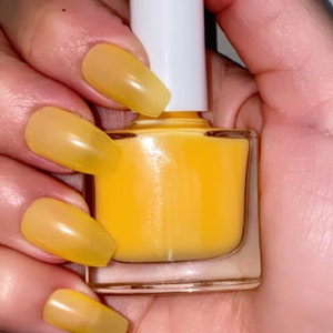 May include: A clear glass bottle of yellow nail polish with a white cap. The bottle is being held by a hand with several nails painted in a matching yellow color.