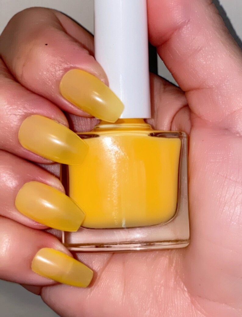 May include: A clear glass bottle of yellow nail polish with a white cap. The bottle is being held by a hand with several nails painted in a matching yellow color.