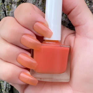May include: A clear glass bottle of orange nail polish with a white cap. The bottle is being held by a hand with orange nail polish on the nails.