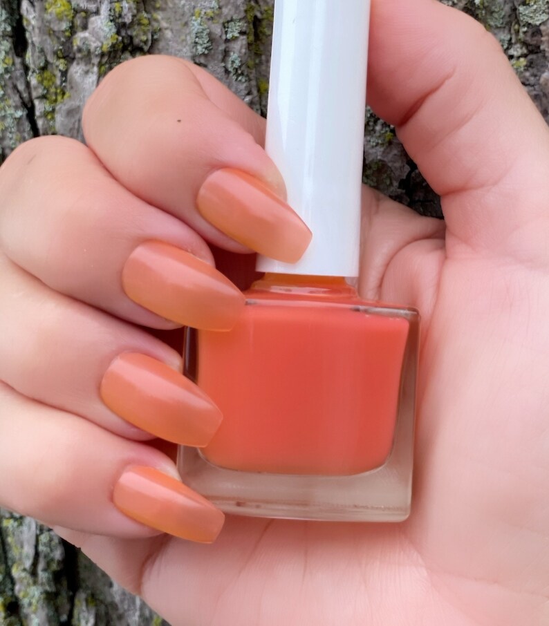 May include: A clear glass bottle of orange nail polish with a white cap. The bottle is being held by a hand with orange nail polish on the nails.