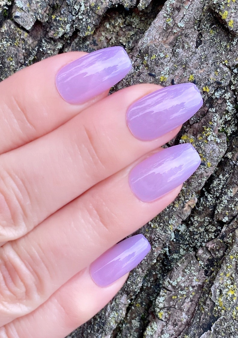 K&ouml;nnte beinhalten: Nahaufnahme einer Hand mit langen, helllila N&auml;geln. Die N&auml;gel sind mit einem gl&auml;nzenden, durchscheinenden Lavendellack lackiert.
