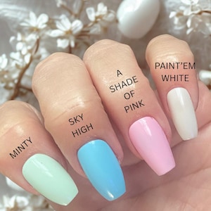 May include: A hand displays five painted fingernails in various pastel colors: mint green, sky blue, light pink, and white. Each nail is labeled with a corresponding color name: "Minty", "Sky High", "A Shade of Pink", and "Paint'em White".