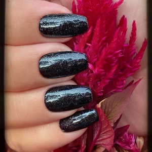 May include: Close-up of four fingernails with dark grey glitter nail polish. The nails are manicured in a square shape and are shown against a background of pink and red flowers.