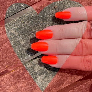 May include: Close-up of a hand with four bright orange nails. The nails are long and have a square shape. The hand is resting on a wooden surface with a heart shape.