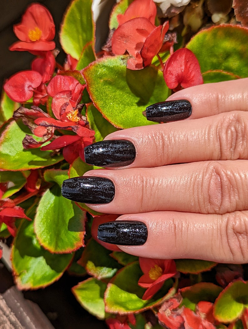 May include: Close-up of a hand with black glitter nail polish on all five nails. The nails are long and have a coffin shape. The hand is resting on a green plant with red flowers.