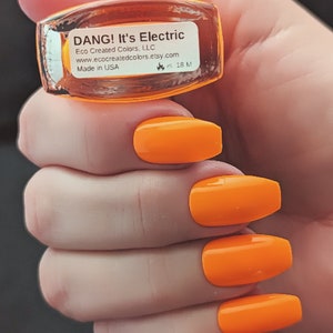 Może przedstawiać: Model dłoni z jaskrawopomarańczowym lakierem do paznokci. Buteleczka lakieru do paznokci jest oznaczona "DANG! It's Electric" i "Eco Created Colors, LLC".