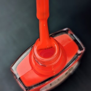 May include: A close-up of a bright orange nail polish bottle with a brush applicator. The bottle is made of clear glass and has a silver cap.
