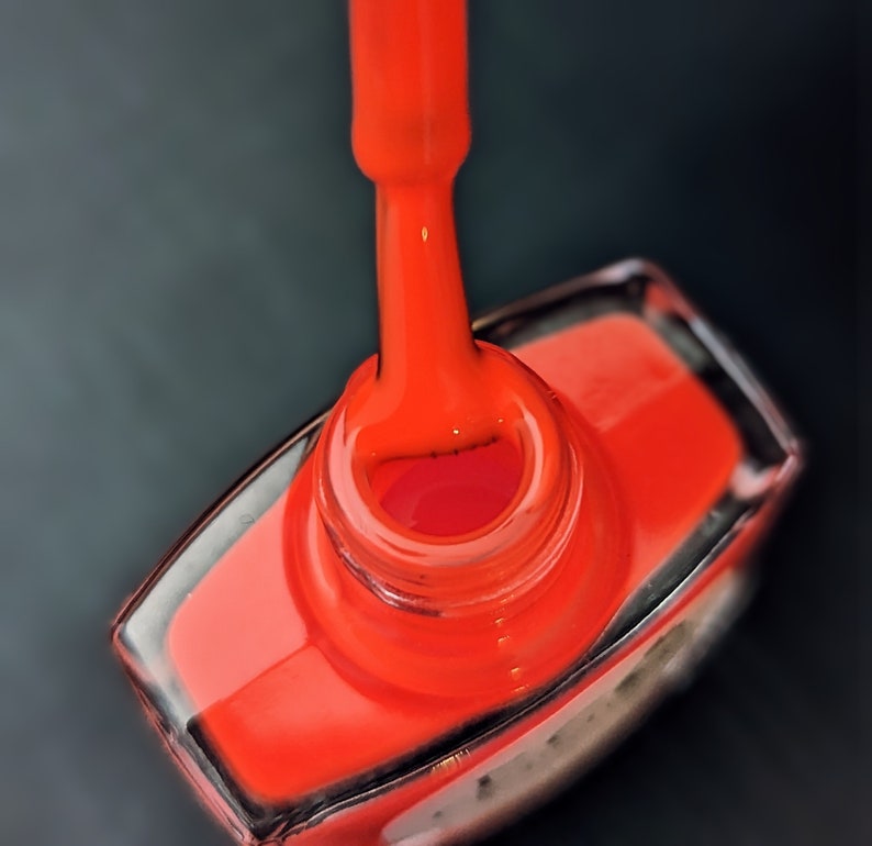 May include: A close-up of a bright orange nail polish bottle with a brush applicator. The bottle is made of clear glass and has a silver cap.