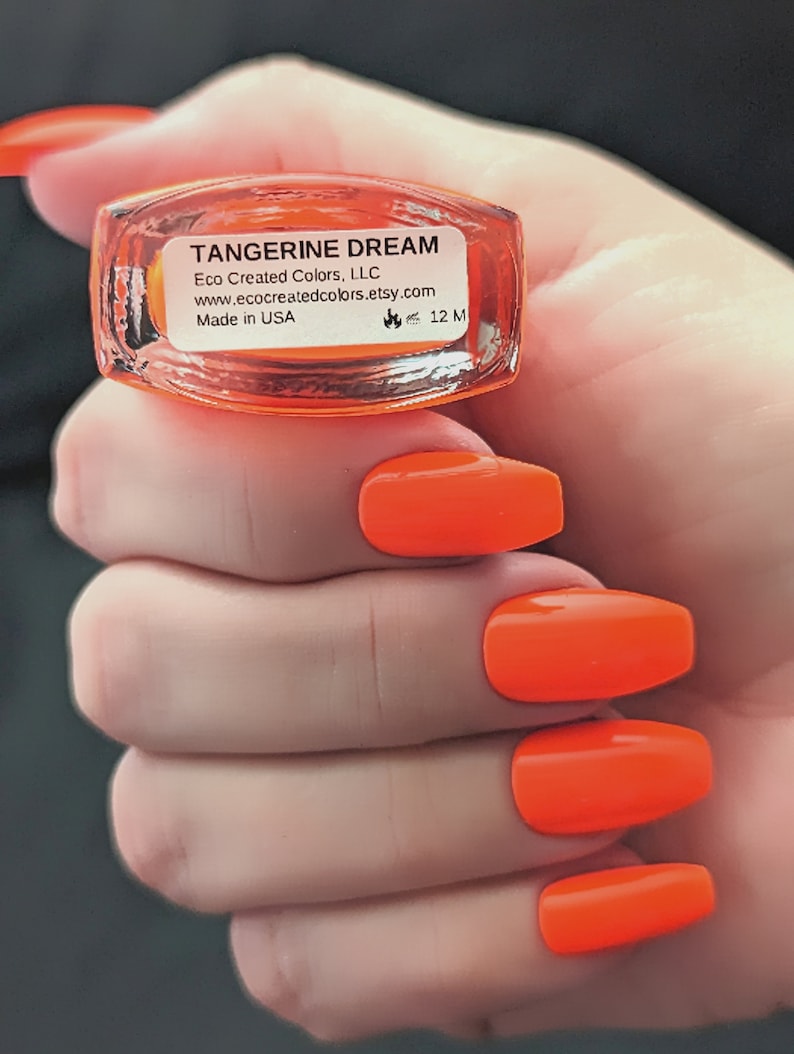 May include: A clear glass bottle of orange nail polish with a black label that reads "TANGERINE DREAM" and "Eco Created Colors, LLC www.ecocreatedcolors.etsy.com Made in USA 12 ML".
