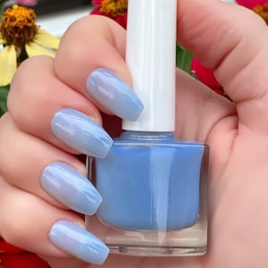 May include: A clear glass bottle of light blue nail polish with a white cap. The bottle is being held by a hand with light blue ombre nails.