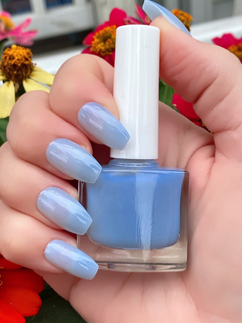 May include: A clear glass bottle of light blue nail polish with a white cap. The bottle is being held by a hand with light blue ombre nails.