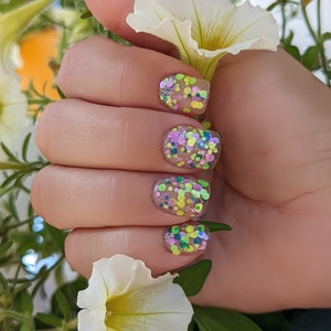 May include: Close-up of a hand with four fingernails painted with a clear base and multi-colored glitter. The glitter is in various sizes and shapes, including circles, squares, and stars. The colors of the glitter are pink, green, yellow, and blue.