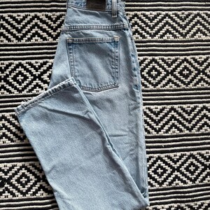 May include: Light blue denim jeans with a classic five-pocket design. The jeans are folded, showcasing the back pocket and the faded wash of the denim. The jeans are laid on a black and white patterned surface.