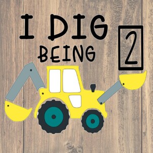 May include: A yellow cartoon excavator with a gray arm and scoop, and the text "I DIG BEING 2" in black. The number 2 is in a black box. The background is a wood grain pattern.