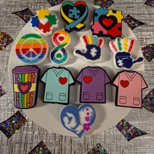 May include: A collection of twelve colorful shoe charms featuring various designs including puzzle pieces, a peace sign, an infinity symbol, handprints, a rainbow coffee cup, three scrub tops in different colors, and a heart-shaped emblem.