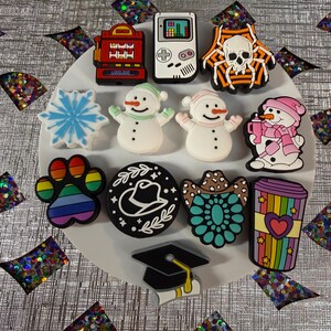 May include: A collection of twelve decorative shoe charms featuring various designs including a red slot machine, a white handheld game console, a skull in an orange spiderweb, a blue snowflake, two snowmen, a rainbow paw print, a cowboy hat, a turquoise concho, a rainbow coffee cup, and a black graduation cap.