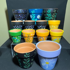 以下が含まれることがあります： さまざまな色とデザインの手描きのテラコッタ植木鉢のコレクション。青、黄、緑、黒の色合いで、花柄や抽象的な模様が施されています。一部の鉢には、自然なテラコッタの縁があります。