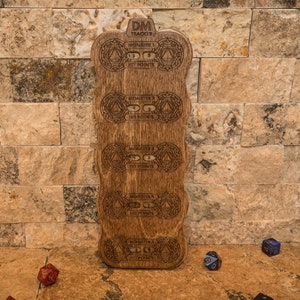 Pode incluir: Um rastreador de DM de madeira com texto e desenhos gravados. O rastreador tem slots para rastrear as estatísticas e os pontos dos monstros. O texto "DM TRACKER" está no topo. Dados espalhados.