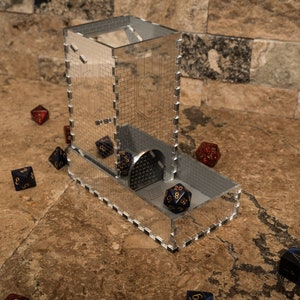 May include: A clear acrylic dice tower with a silver metallic finish. The tower has a slot for dice to roll out of and a small compartment at the bottom to catch the dice. There are several dice scattered around the tower, some are blue and some are red.
