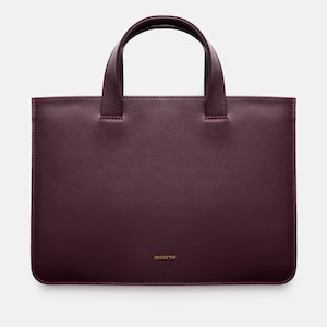 Könnte beinhalten: Eine tiefburgunderrote Ledertasche mit rechteckiger Silhouette, zwei Tragegriffen und abgerundeten Ecken. Das Wort "incarne" ist in kleiner goldener Schrift nahe der unteren Mitte der Vorderseite eingeprägt und hat eine glatte Oberfläche.