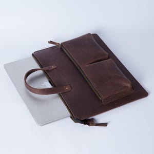 Könnte beinhalten: Eine braune Laptop-Hülle aus Leder mit Griff und zwei Vordertaschen. Die Hülle hat einen Reißverschluss und ein silberner Laptop ist teilweise sichtbar. Die Hülle dient zum Schutz und Transport eines Laptops.