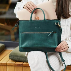 Könnte beinhalten: Eine dunkelgrüne Laptoptasche aus Leder mit zwei Tragegriffen, einem abnehmbaren Schulterriemen und einer Reißverschlusstasche auf der Vorderseite. Die Tasche ist mit den Initialen G.S. geprägt und enthält einen hellen Laptop im Hauptfach.