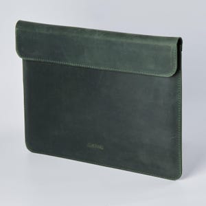 Könnte beinhalten: Eine dunkelgrüne Laptop-Hülle aus Leder mit Klappenverschluss. Die Hülle hat eine strukturierte Oberfläche und sichtbare Nähte an den Kanten. Der Markenname "INCARNE" ist dezent auf der unteren Vorderseite geprägt.