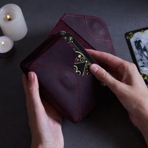 May include: A burgundy leather card holder with a flap that opens to reveal a black and gold tarot card. The card holder is made of high-quality leather and has a stitched design.