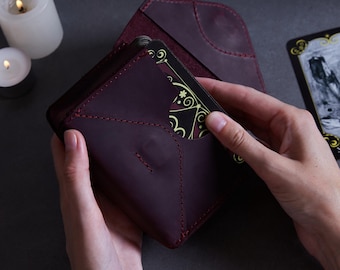 May include: A burgundy leather card holder with a flap that opens to reveal a black and gold tarot card. The card holder is made of high-quality leather and has a stitched design.