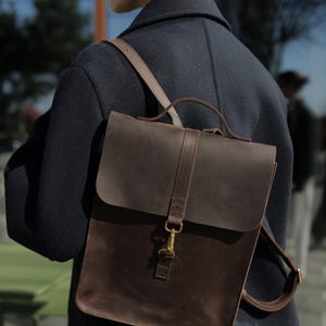 Könnte beinhalten: Ein dunkelbrauner Lederrucksack mit Überschlagklappe, Tragegriff und verstellbaren Schultergurten. Die Tasche wird durch einen mittigen, vertikalen Lederriemen und einen messingfarbenen Metallhakenverschluss an der Vorderseite gesichert.