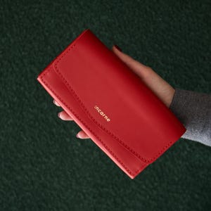 Pode incluir: Uma carteira retangular de couro vermelho com fecho de aba e costuras visíveis ao longo das bordas. O nome da marca :incarne está gravado em letras douradas pequenas na aba frontal da carteira, que possui um acabamento liso e fosco.