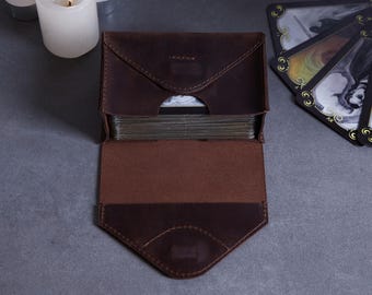 May include: A dark brown leather tarot card case with a fold-over envelope flap and visible stitching. The case is open, revealing a deck of cards inside. The leather has a matte finish and a structured rectangular shape with a pointed flap closure.