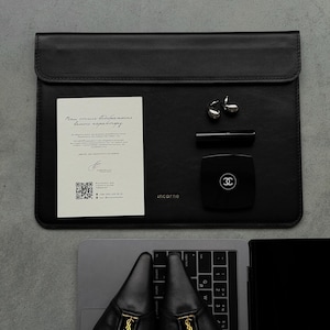 Könnte beinhalten: Eine schwarze Laptoptasche aus Leder mit dem eingeprägten Markennamen "incarne" auf der Vorderseite. Darauf liegen eine weiße Informationskarte mit Text und QR-Code, zwei silberfarbene Ohrringe, ein schwarzer Lippenstift und ein schwarzes Puderetui.