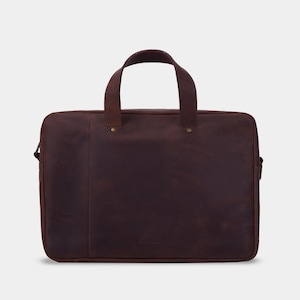May include: A dark brown leather laptop bag with two top handles, a rectangular silhouette, and a vertical seam detail on the front. The bag features brass-coloured rivets at the base of the handles and a small embossed logo at the bottom centre.