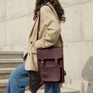 May include: A dark burgundy leather satchel with a rectangular flap, a front pocket, and brass-coloured metal hardware. The bag features a long shoulder strap and a structured vertical design, worn over a beige blazer and blue denim jeans.