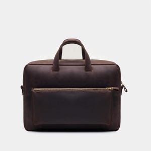 Könnte beinhalten: Eine dunkelbraune Aktentasche aus Leder mit rechteckiger Silhouette, zwei Tragegriffen und einer horizontalen Vordertasche mit messingfarbenem Reißverschluss. Die Tasche verfügt über ein Hauptfach mit Reißverschluss und eine matte Oberfläche.