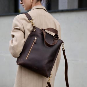 May include: A dark brown leather tote bag with a gold zipper and hardware. The bag has a shoulder strap and a top handle. The bag is worn over the shoulder. The person is wearing a tan blazer.