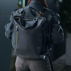 Puede incluir: Una mochila negra con acabado liso, que presenta un bolsillo vertical con cremallera en la parte frontal, herrajes en tono dorado y dos asas superiores. La bolsa incluye correas ajustables y un pequeño logotipo dorado impreso cerca del borde inferior.