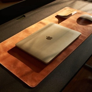 May include: A tan leather desk mat with a stitched edge, a silver laptop, a white mouse, and a small notepad. The laptop features an Apple logo. The desk is dark grey.