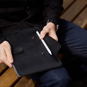 Pode incluir: Uma capa de tablet de couro preto com uma caneta branca. A capa tem um fecho de pressão e rebites cor de latão. A caneta é inserida em um bolso dentro da capa. A capa está nas mãos de uma pessoa.